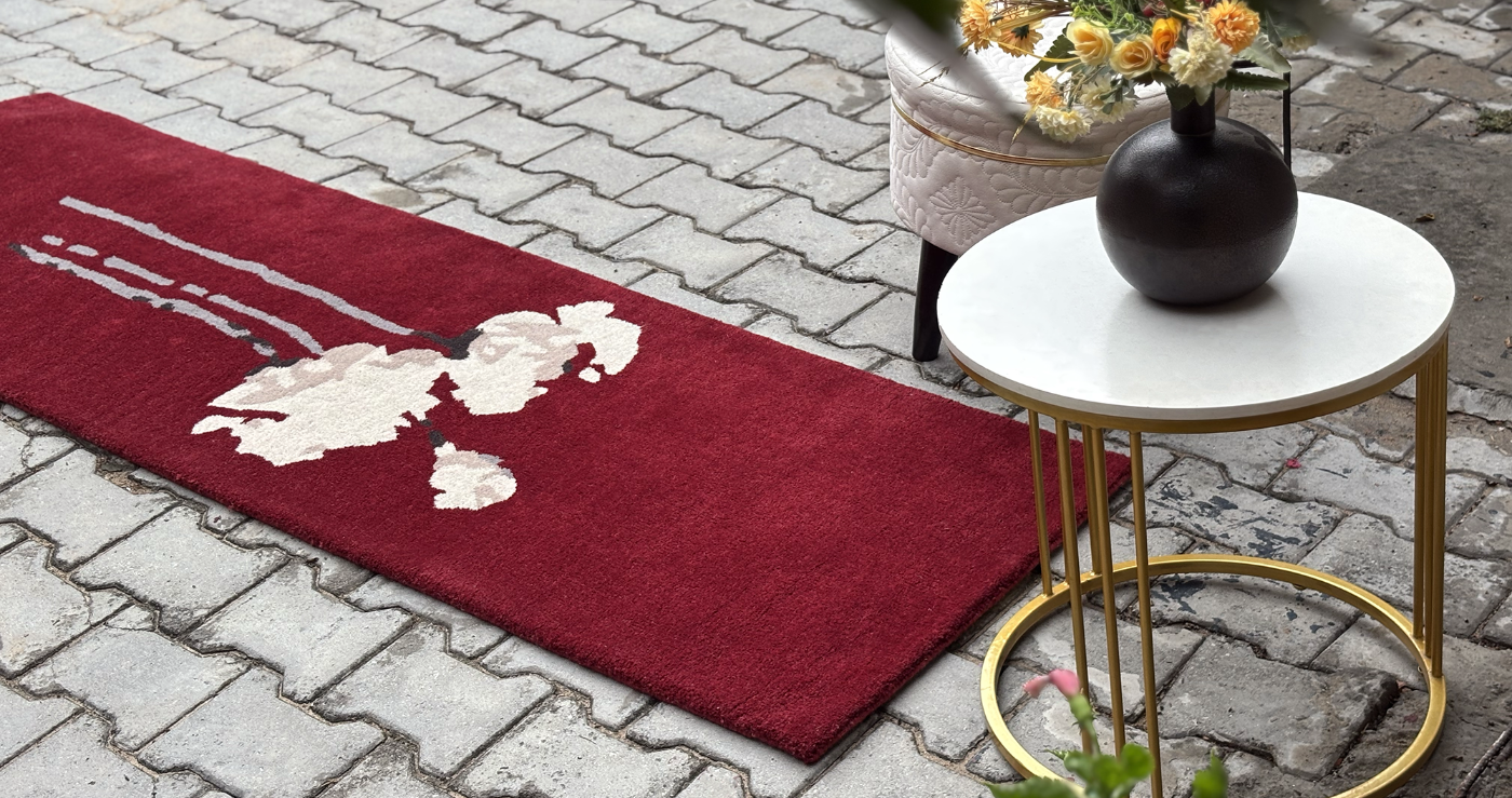Red rug with white floral design on a stone pavement, next to a small round table with a black vase and flowers.